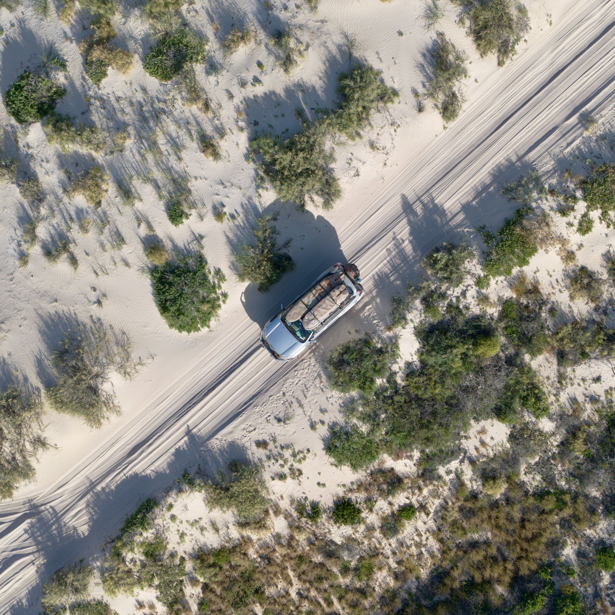 A 4WD driving through sand dunes with green shrubs on the side, photographed from above by a drone.